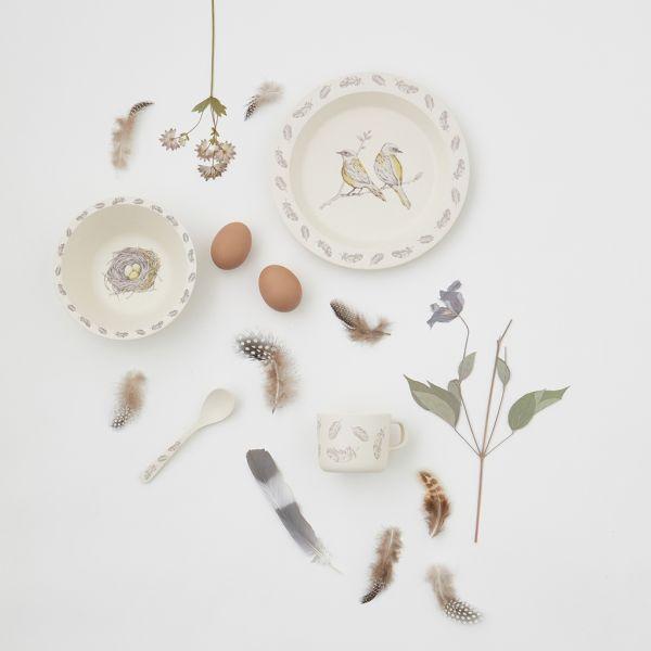 Set pappa decorato con immagini di uccellini (1 fondina, 1 piatto piano, 1 tazza con manico e 1 cucchiaio) su una tavola con altri oggetti