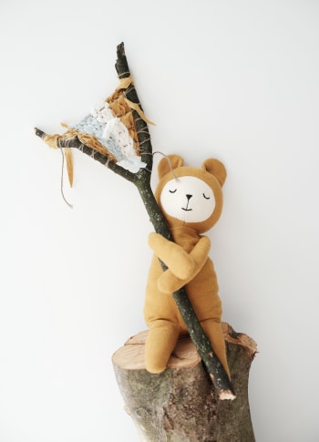 Orsacchiotto in morbido cotone color mostarda abbracciato ad un ramo sopra un ceppo