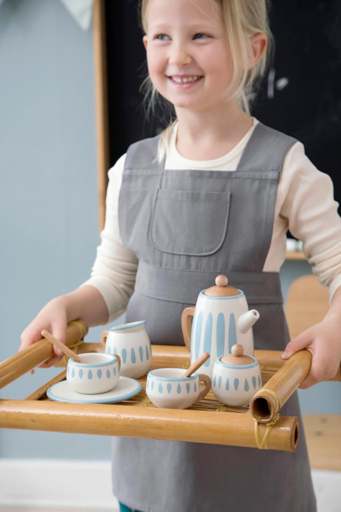 Bambina con vassoio con su set da tè comprendente una teiera con coperchio, una lattiera, una zuccheriera con coperchio, due tazze da tè con piattini e due cucchiaini da tè, tutti realizzati in legno.