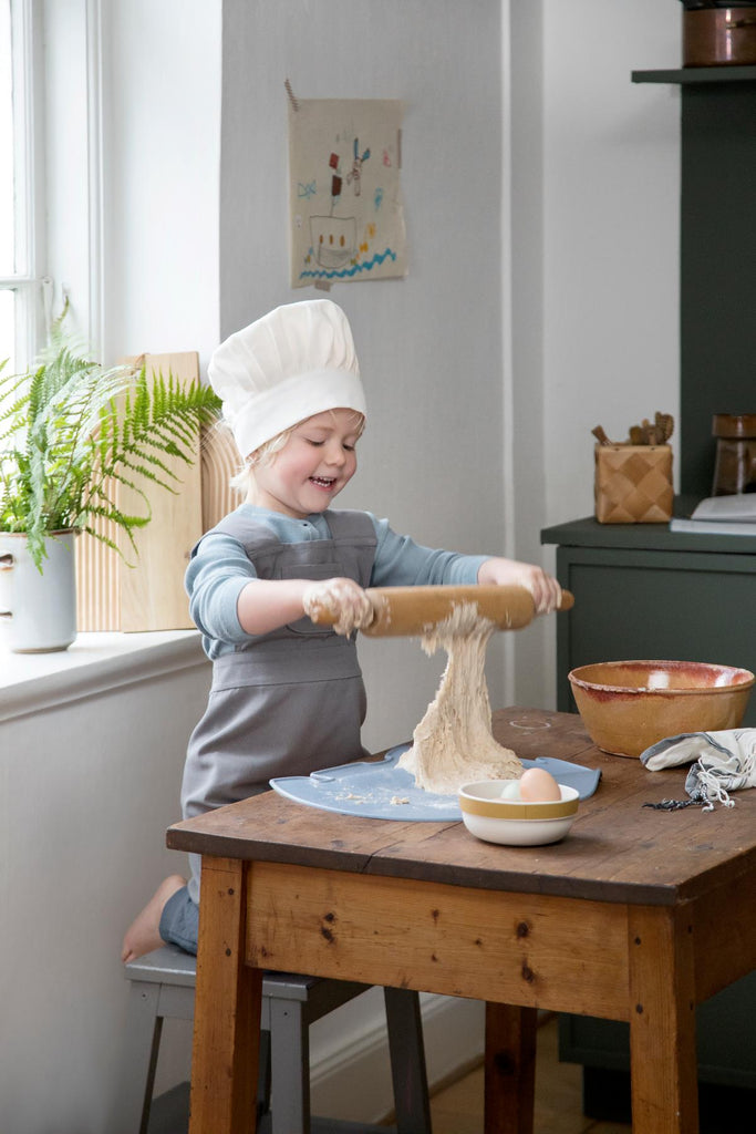 Bmbina con grenbiulino da cucina grigio e cappello da cuoco bianco intenta a lavorare la pasta con un mattarello