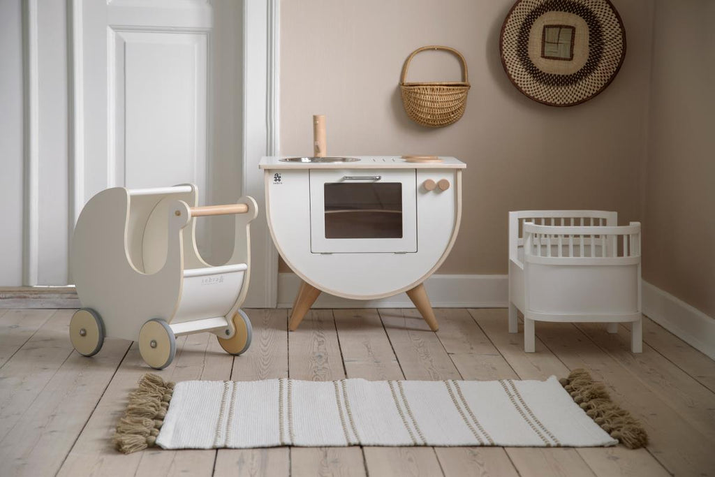 Mini cucina in legno color bianco con forno, lavello e due fuochi con a fianco una carrizina e un lettino bianchi
