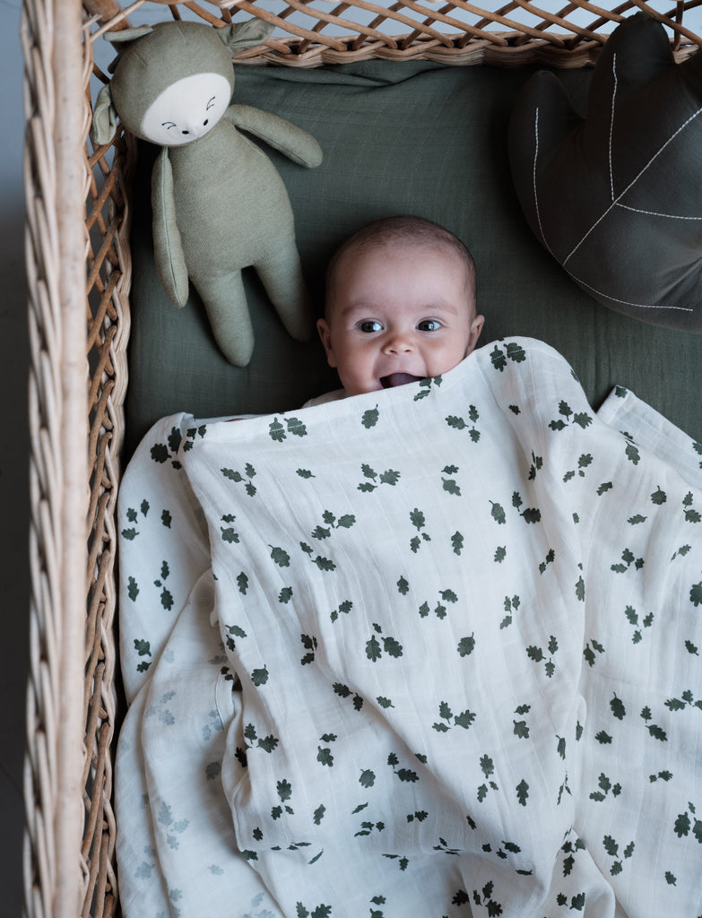 Peluche verde oliva a forma di draghetto addormentato in una cesta accanto ad un neonato con copripiumino a fiori