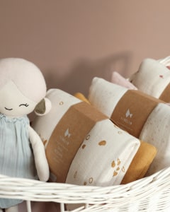 Bambola di  pezza Apple in un cesto accanto a confezioni di biancheria da letto per bambole